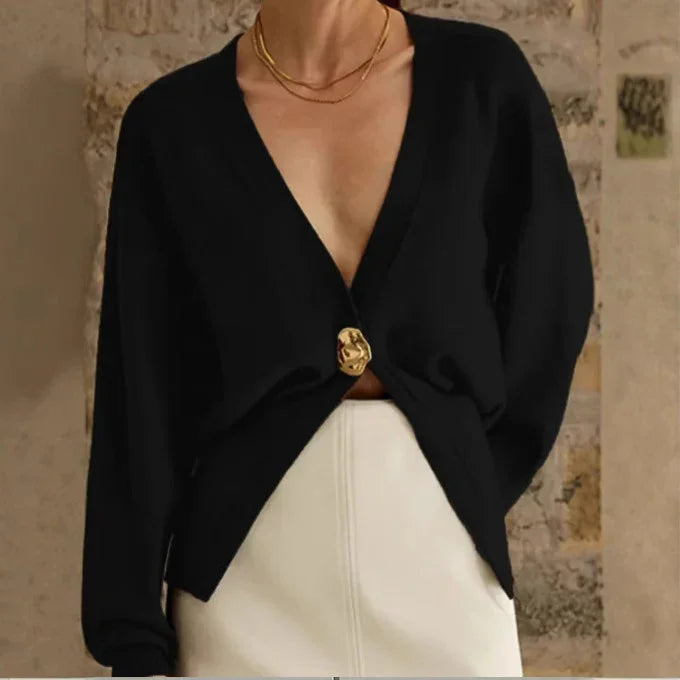 Woman wearing a black deep V-neck cardigan with gold button and white skirt against a stone wall background
