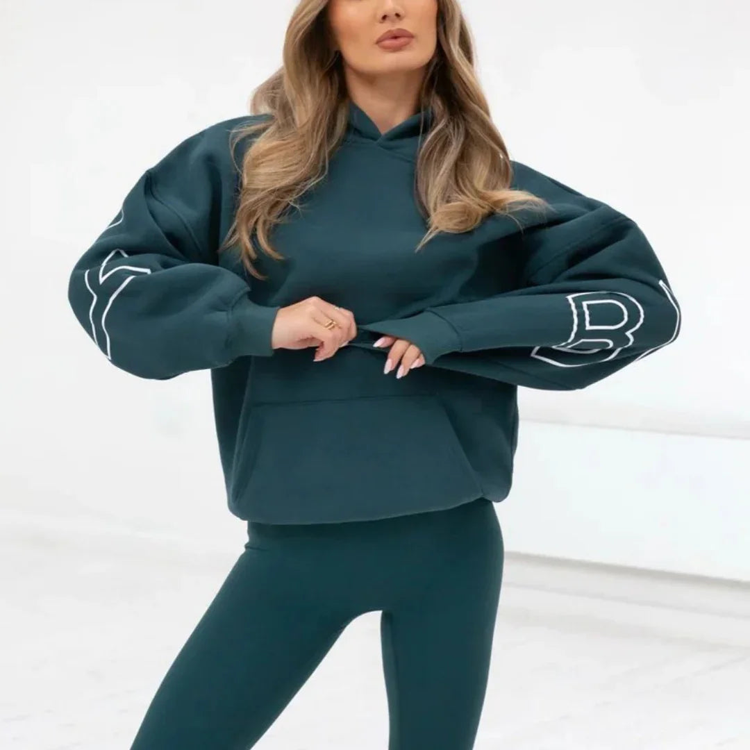 Woman wearing dark teal oversized hoodie with graphic sleeves and matching leggings in minimalist studio
