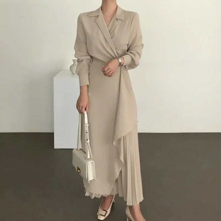 Woman wearing beige wrap dress with pleated skirt detail, holding white handbag, standing indoors