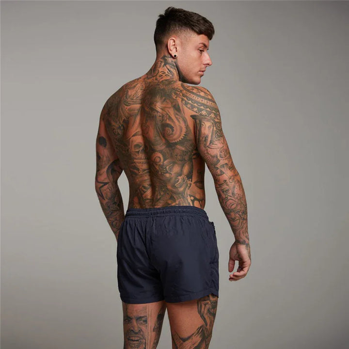 Muscular man with full tattoo sleeve and back tattoos wearing navy blue shorts against gray background
