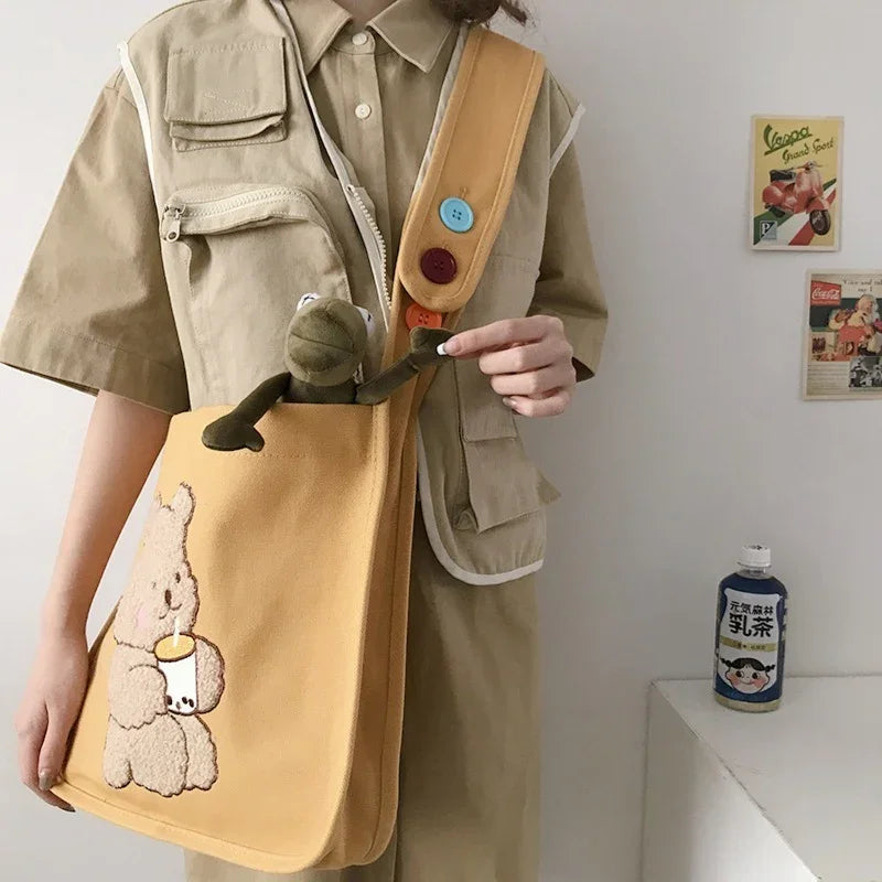 Person wearing beige utility shirt holding yellow shoulder bag with teddy bear and bubble tea design and stuffed frog toy