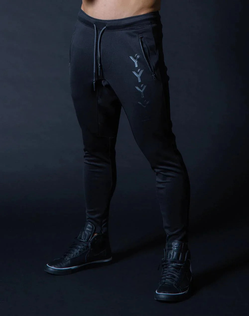 Man wearing black slim-fit jogger pants with front zipper pockets and black sneakers against dark background
