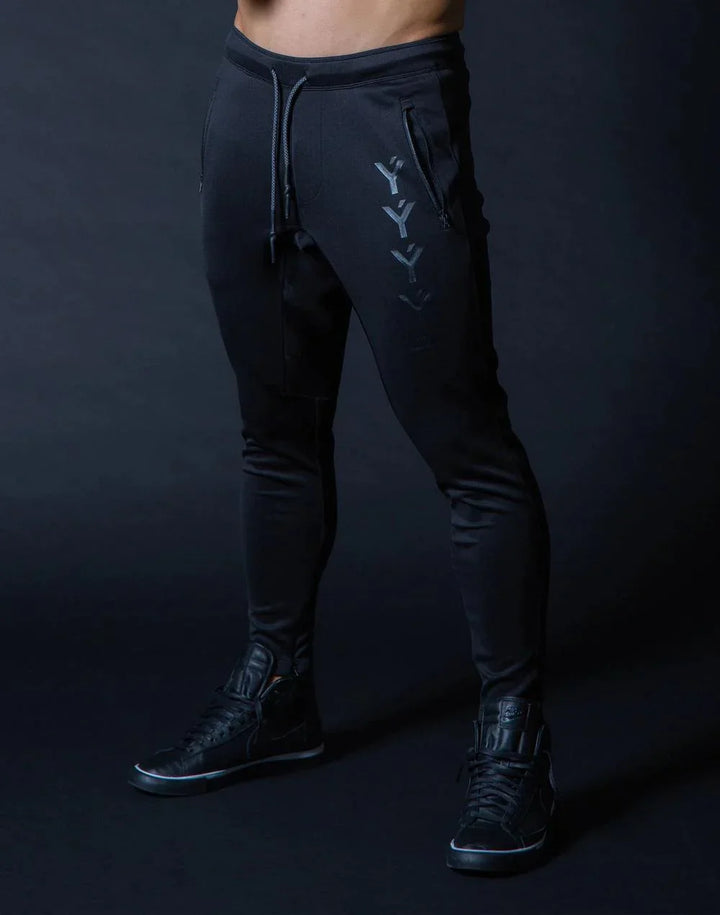 Man wearing black slim-fit jogger pants with front zipper pockets and black sneakers against dark background