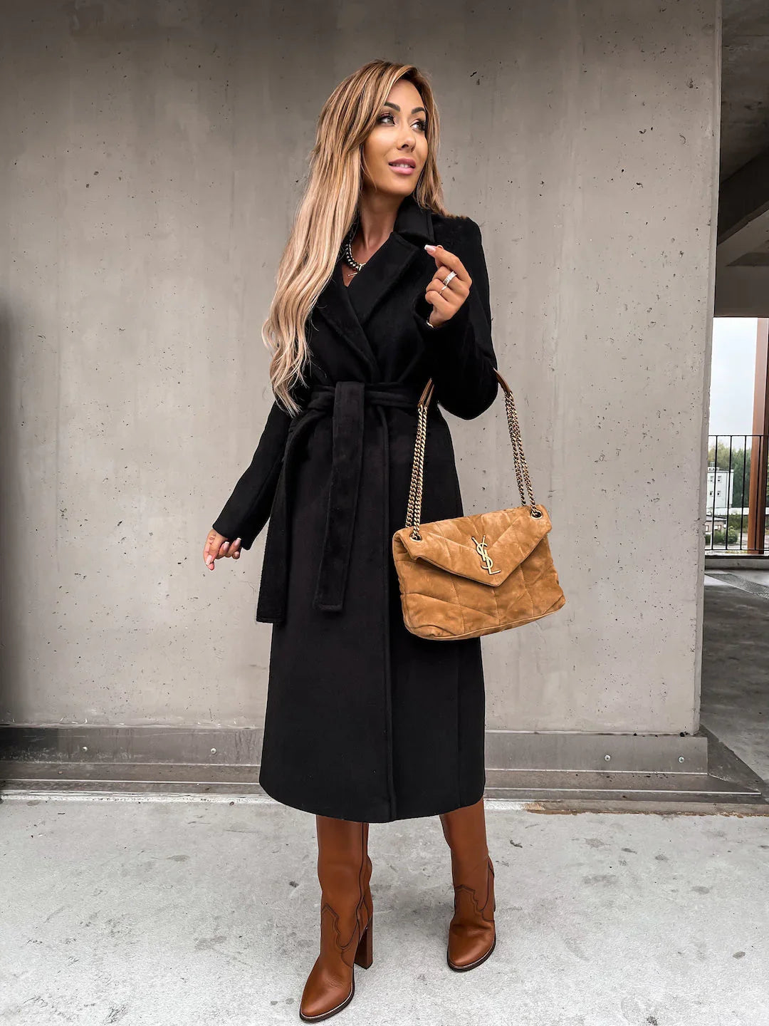 Woman wearing a black belted coat, brown leather boots, and carrying a tan suede YSL handbag against a concrete background