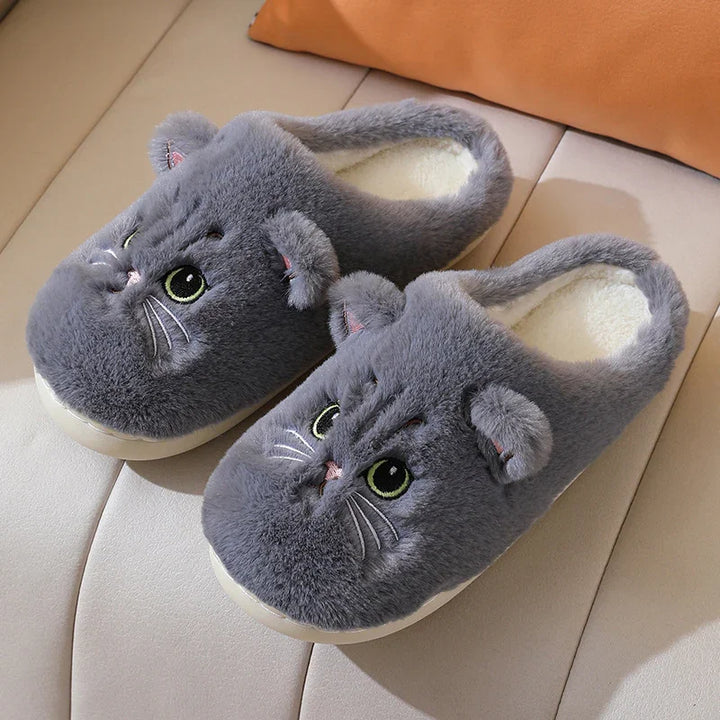 Gray plush slippers with cute cat faces, ears, and embroidered green eyes on beige sofa