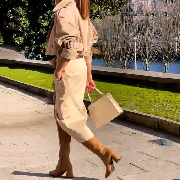 Woman walking outdoors wearing beige trench coat, midi skirt, matching boots, and carrying beige handbag