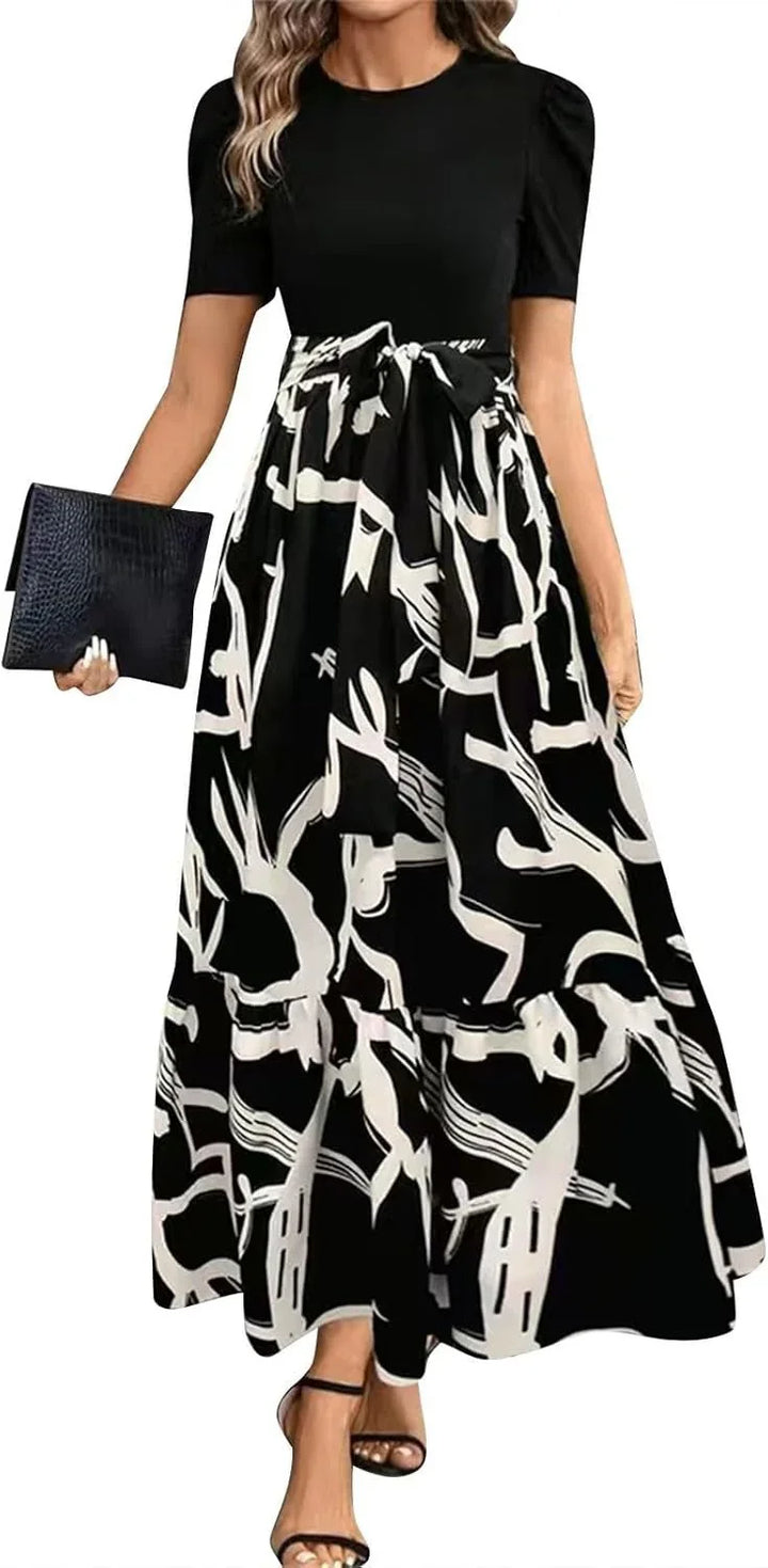 Woman wearing black top with short sleeves and black-and-white abstract print maxi skirt with bow belt, holding black clutch