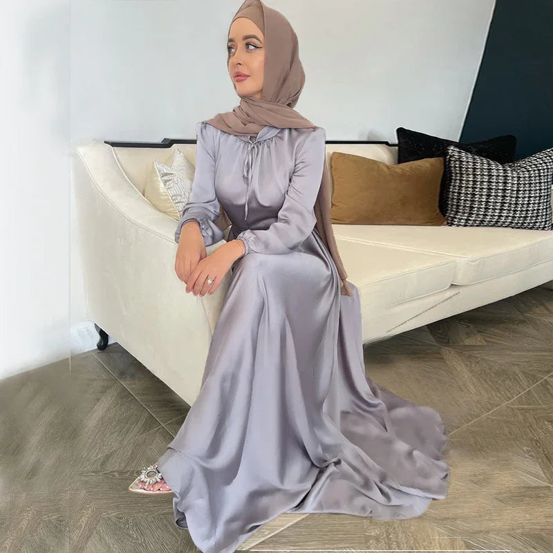 Woman wearing a silky lavender modest dress and taupe hijab sitting on beige sofa with decorative pillows