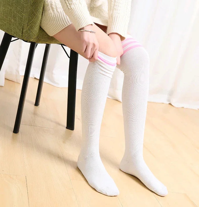Person sitting on green chair wearing white knit sweater and pulling on white knee-high socks with pink stripes