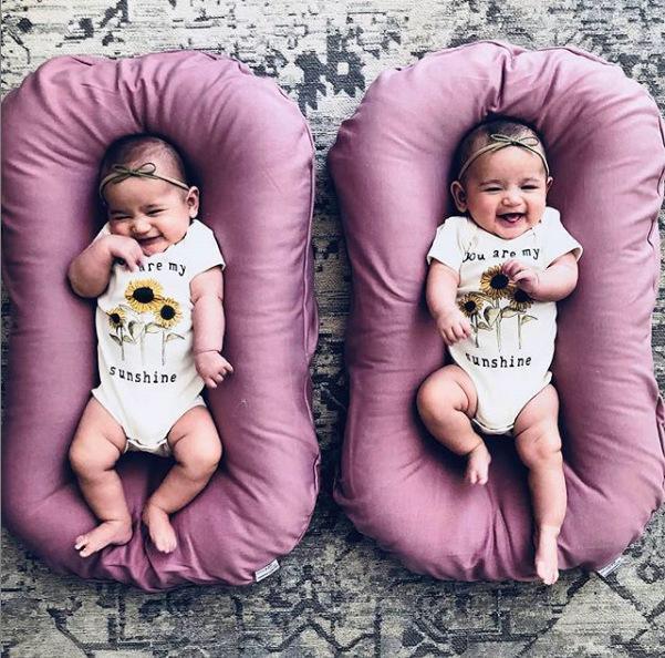 Two smiling babies wearing matching sunflower print onesies and headbands lying on purple cushioned loungers