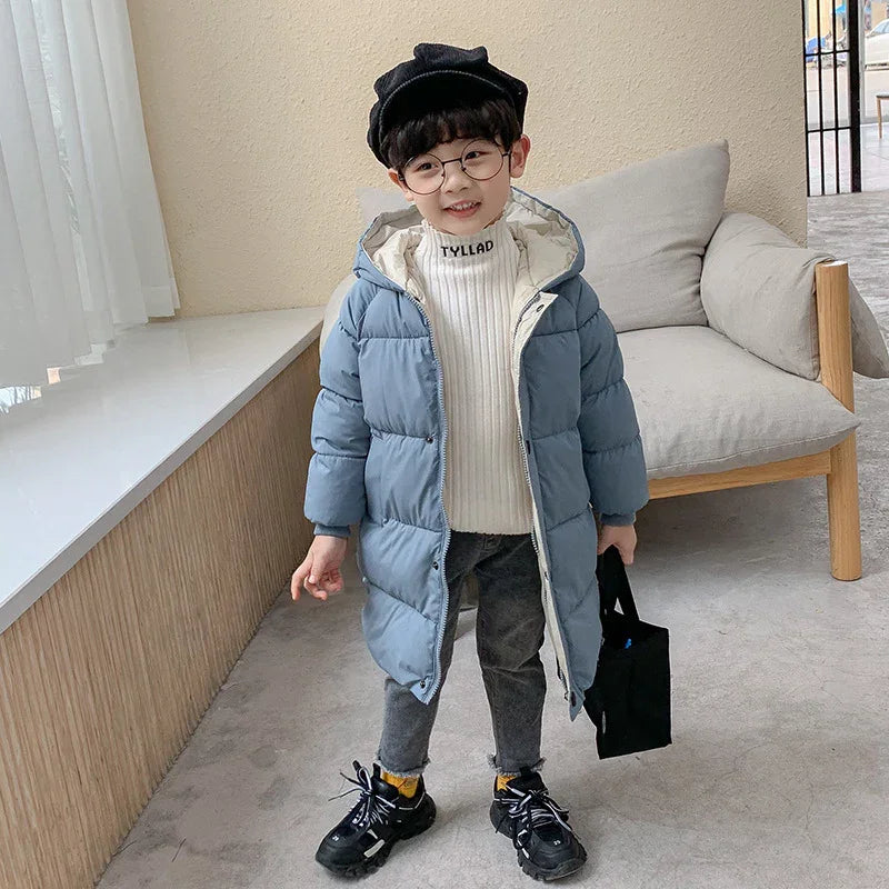 Cute little boy wearing black hat, round glasses, blue puffer coat, white turtleneck sweater, gray pants, and black sneakers indoors