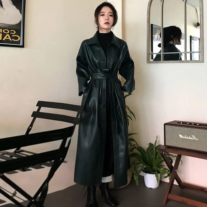Woman wearing a long black leather belted coat and black boots standing indoors with minimalistic decor