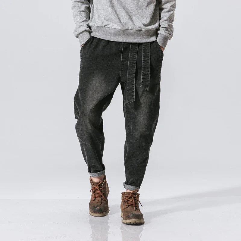 Man wearing black baggy pants with belt detail, grey sweatshirt, and brown boots on white background