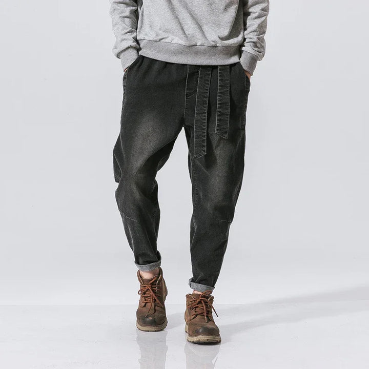 Man wearing black baggy pants with belt detail, grey sweatshirt, and brown boots on white background