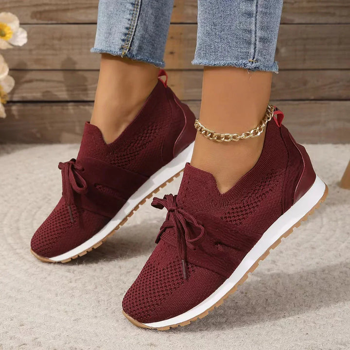 Woman wearing burgundy knitted wedge sneakers with white soles and gold ankle chain on beige carpet