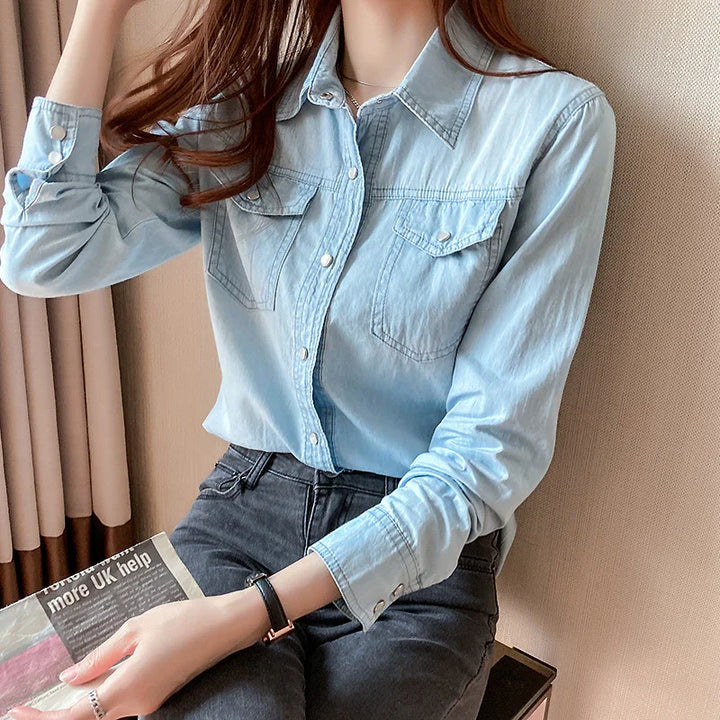 Woman wearing light blue denim shirt and dark jeans sitting on beige chair holding a newspaper