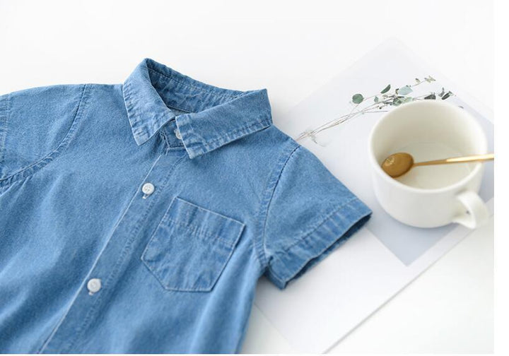 Light blue denim short-sleeve shirt with front pocket next to white cup with gold spoon on minimal white background