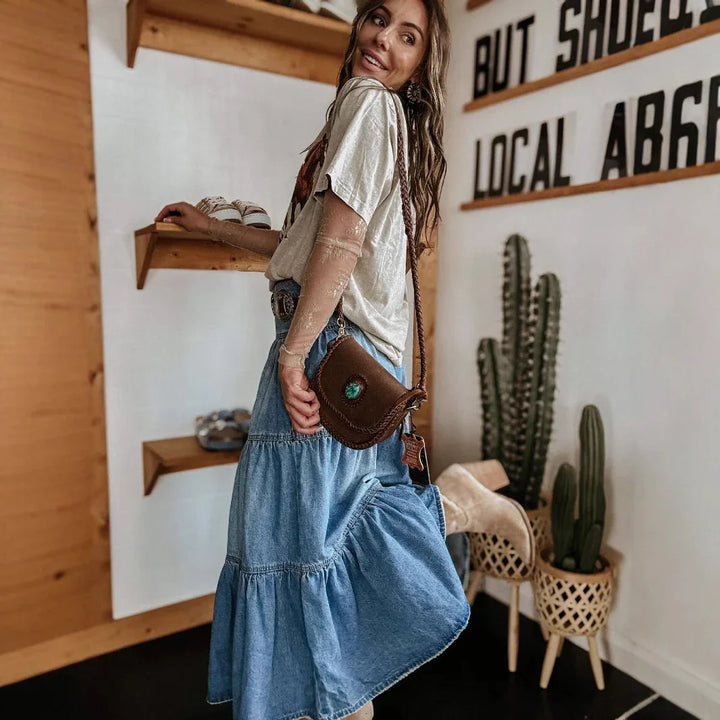 Smiling woman in a beige top and blue denim skirt posing indoors with leather crossbody bag and cactus plants