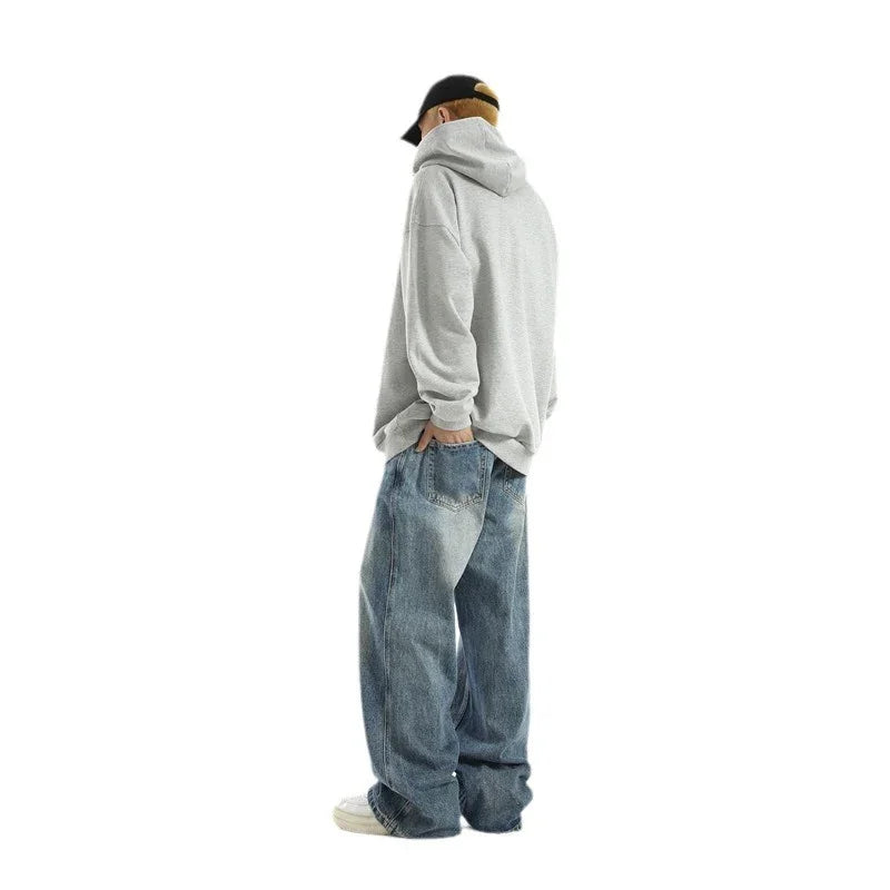 Man wearing a gray hoodie, black cap, and loose blue jeans standing sideways on white background