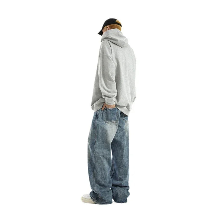 Man wearing a gray hoodie, black cap, and loose blue jeans standing sideways on white background