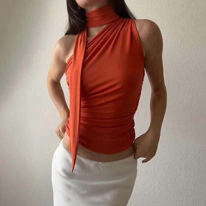 Woman wearing rust orange sleeveless wrap top with scarf detail paired with white skirt against plain wall