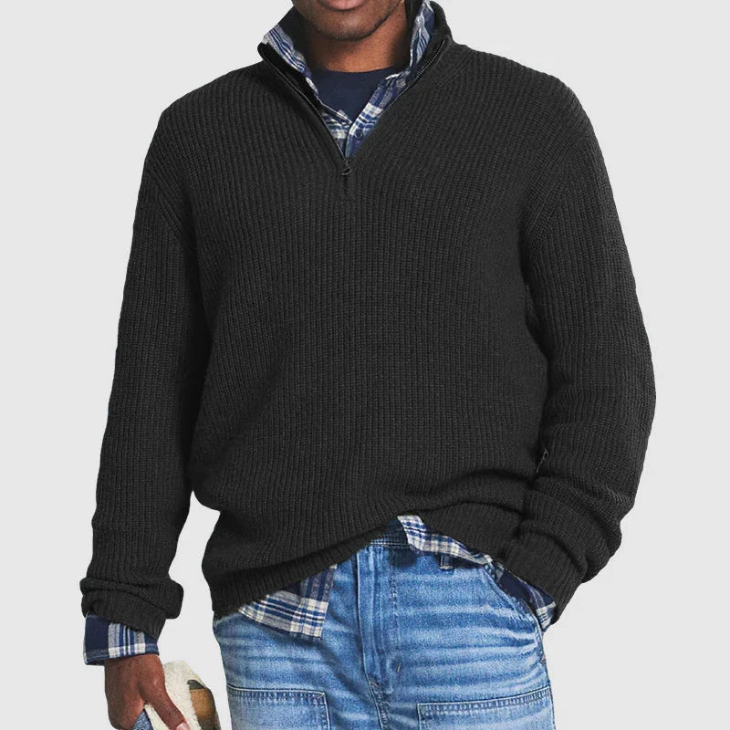 Man wearing black half-zip knit sweater over blue plaid shirt with light blue jeans against white background