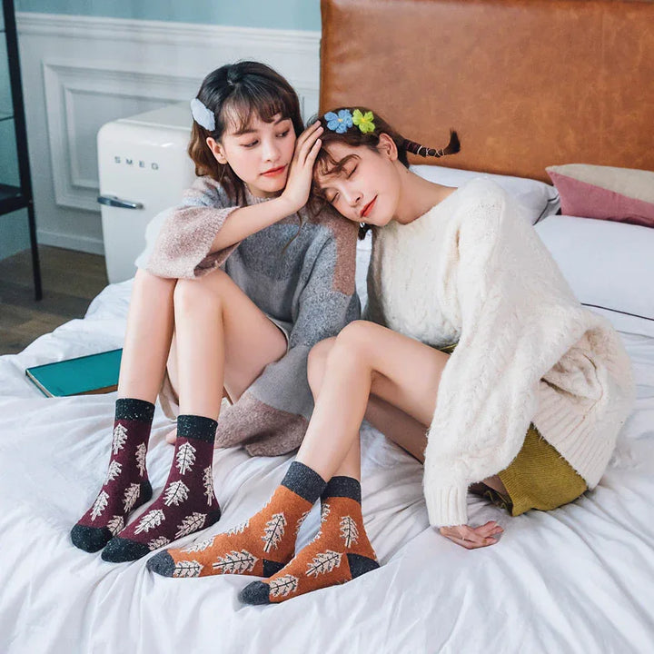 Two women sitting on bed wearing cozy sweaters and colorful leaf-patterned socks, in a modern bedroom