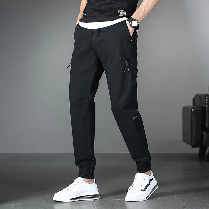 Man wearing black cargo jogger pants and white sneakers standing indoors on gray floor