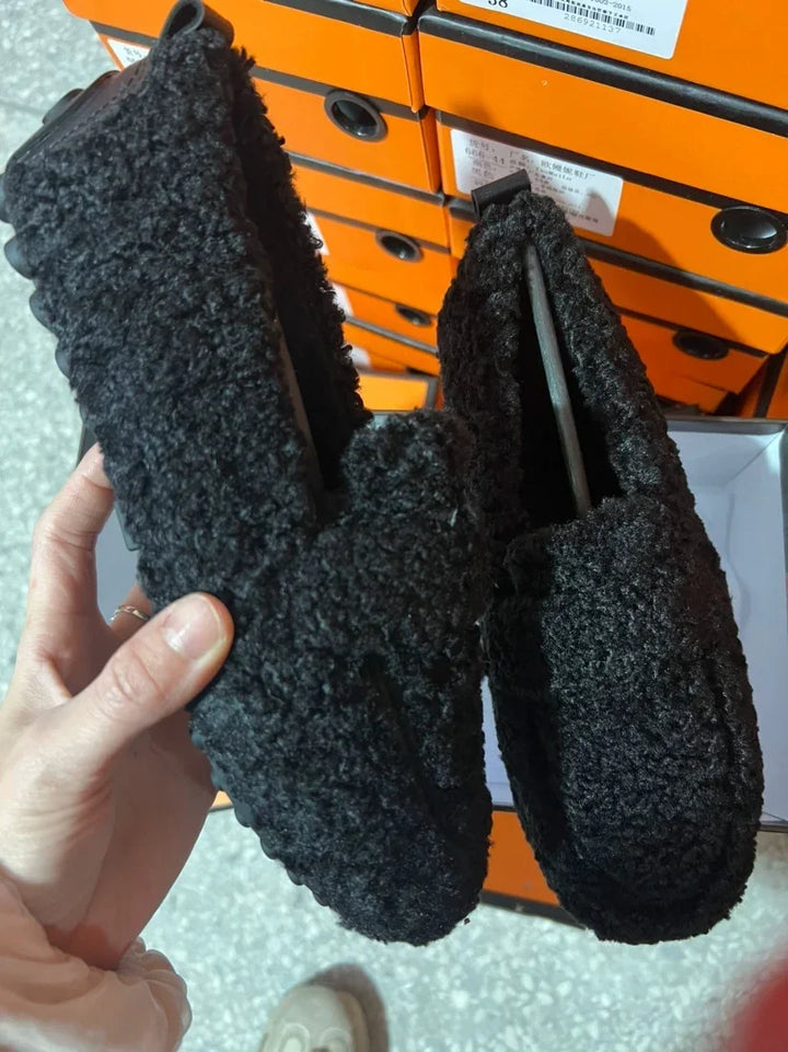 Hand holding a pair of black fuzzy slip-on shoes with textured soles in front of stacked orange shoeboxes