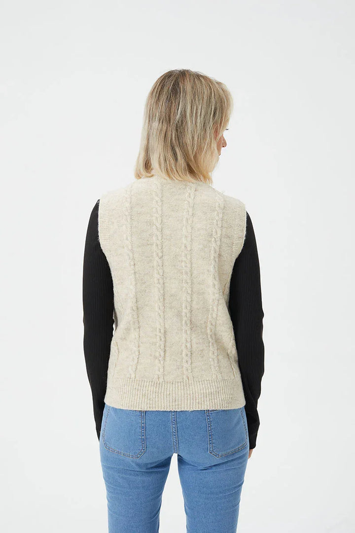 Woman wearing a cream cable knit sleeveless sweater over a black long-sleeve top with blue jeans, back view