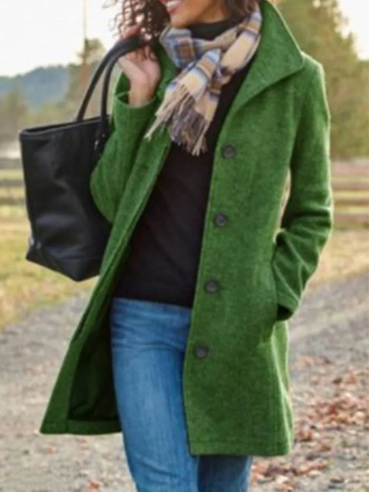 Woman wearing a green buttoned coat, plaid scarf, blue jeans, and carrying a black tote bag outdoors