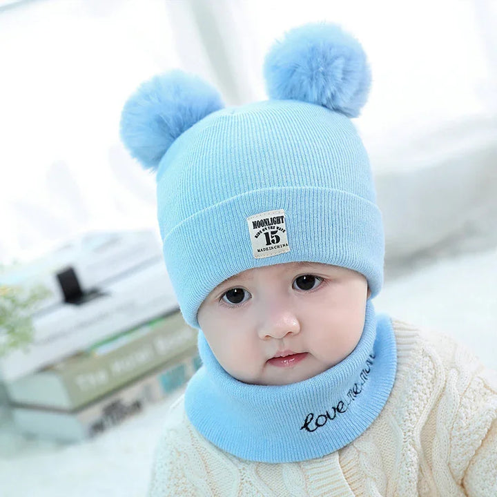 Baby wearing light blue knit hat with pom-poms and matching neck warmer indoors