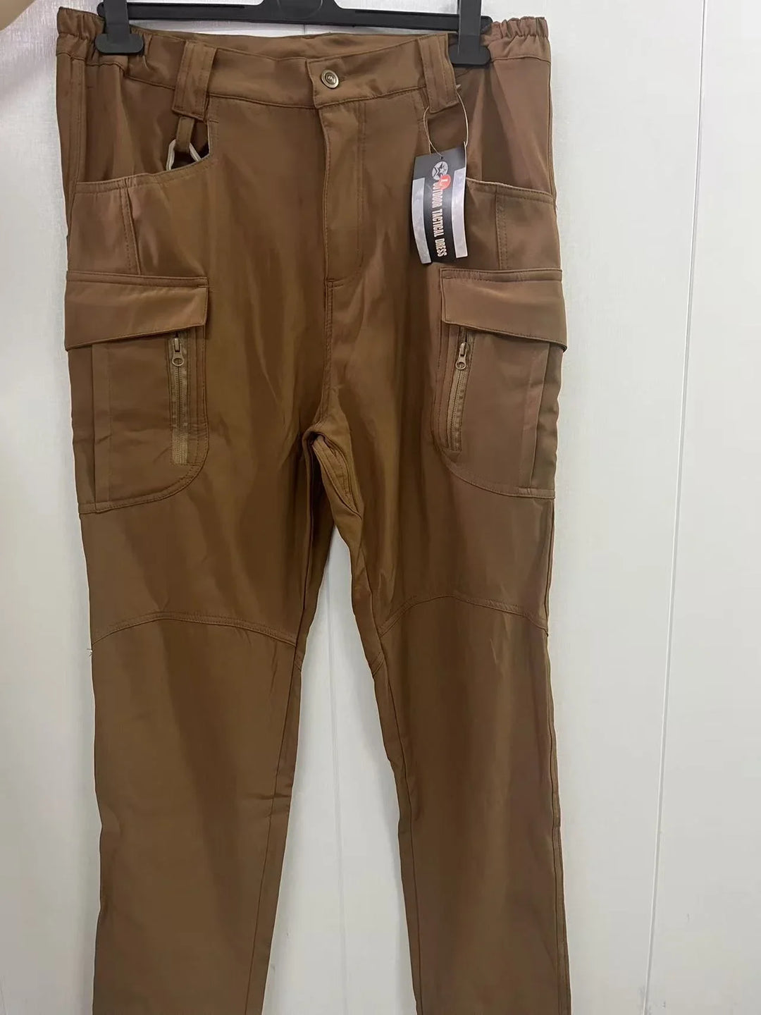 Brown tactical cargo pants with multiple zippered pockets hanging on a hanger against a white wall