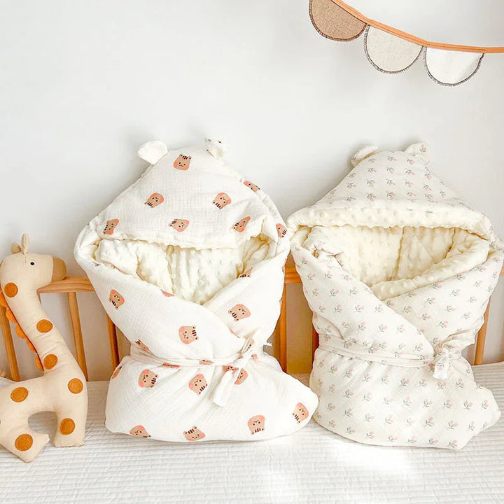 Two cozy baby swaddle blankets with bear ears, one with teddy bear print, the other with floral design, on crib