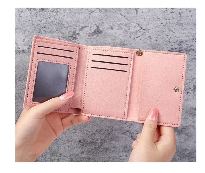 Hands holding open pink trifold leather wallet with card slots and clear ID window on grey background