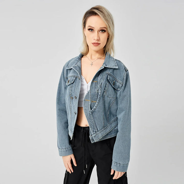 Blonde woman wearing blue denim jacket, white crop top, and black pants against gray background