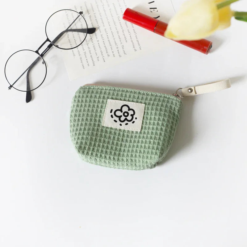 Green waffle-textured fabric pouch with flower patch, round glasses, red lipstick, and book on white surface