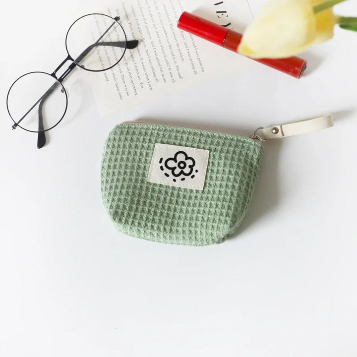 Green waffle-textured fabric pouch with flower patch, round glasses, red lipstick, and book on white surface
