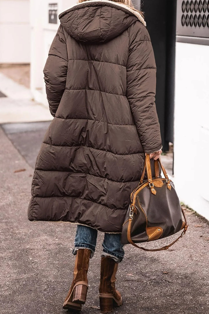 Woman wearing long brown puffer coat with hood, blue jeans, brown heeled boots, carrying dark brown leather handbag
