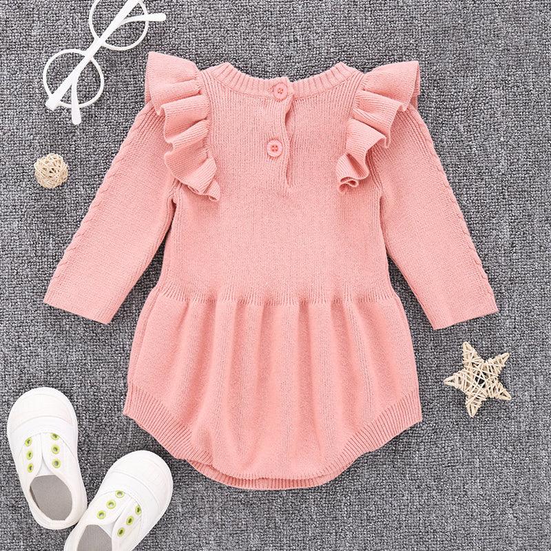 pink knitted baby dress with ruffles on shoulders, white baby shoes, white glasses on grey fabric background