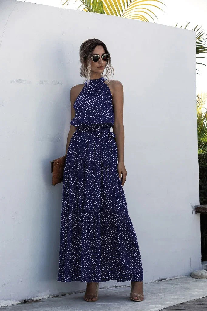 Woman wearing a navy blue halter maxi dress with white polka dots, sunglasses, and holding a brown clutch.
