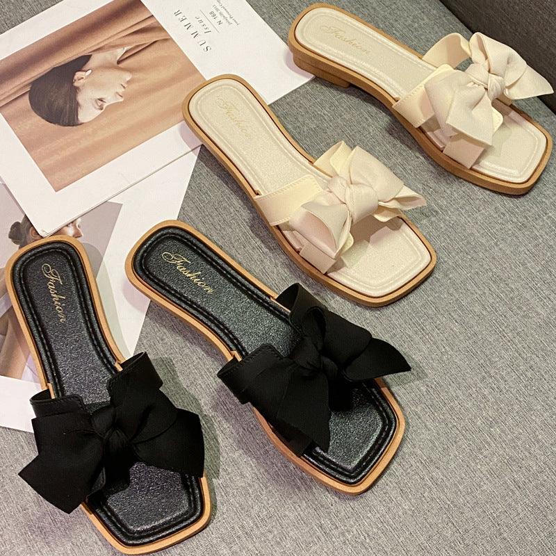 Black and beige flat slide sandals with large fabric bows on gray surface near fashion magazine pages