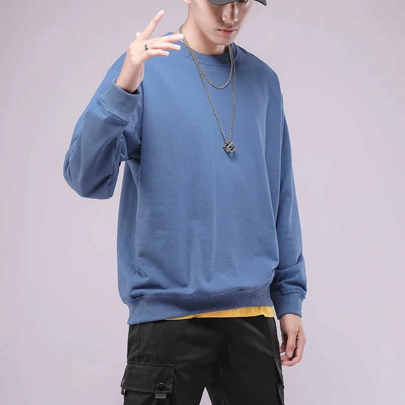 Man wearing blue sweatshirt, black cargo pants, black cap, and cube necklace on plain background