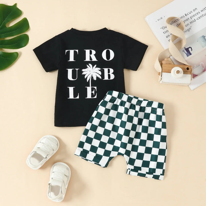 Toddler outfit with black 'TROUBLE' t-shirt and green-white checkered shorts on beige background