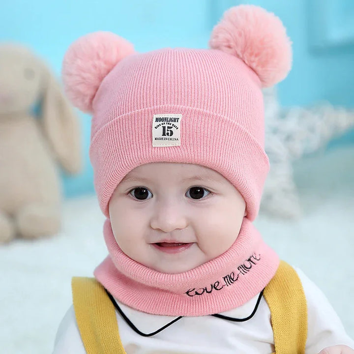 Cute baby wearing pink knit hat with pom-poms and matching neck warmer in cozy indoor setting