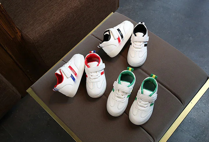 White toddler sneakers with red, black, and green accents on brown cushioned bench