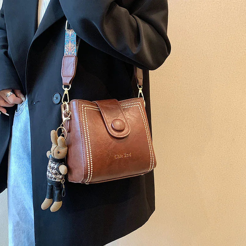 Brown leather crossbody bag with decorative stitching and stuffed animal keychain, worn with black coat and jeans