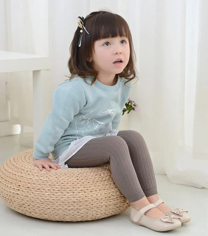 Young girl sitting on woven pouf wearing blue sweater, brown leggings, and beige shoes indoors