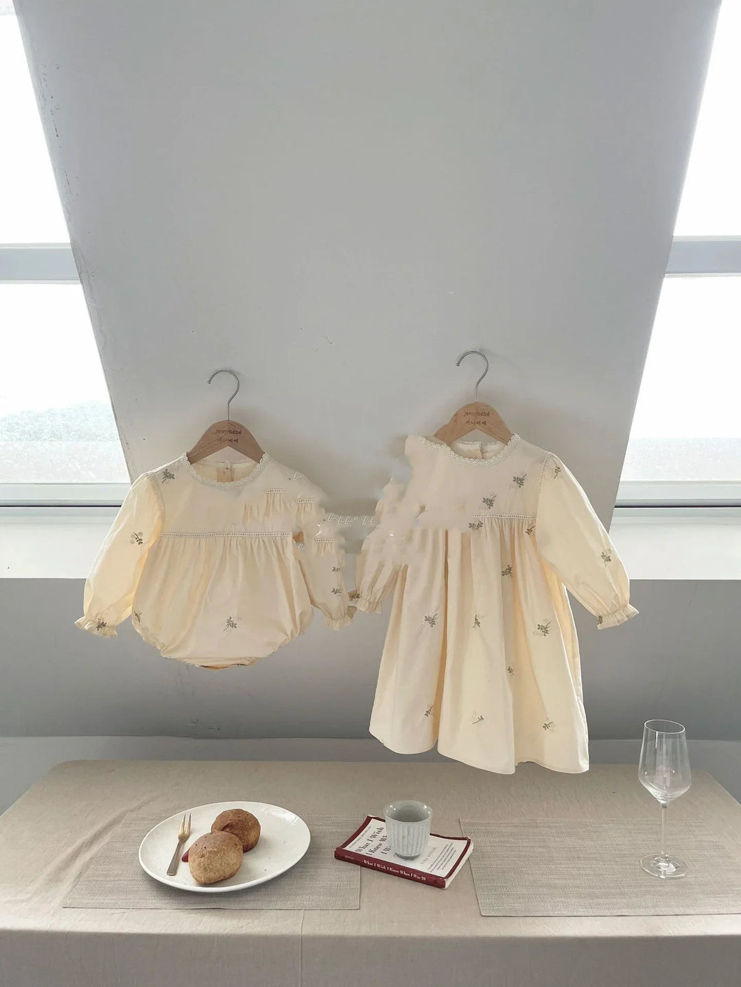 Two cream-colored baby dresses with floral embroidery hanging on wooden hangers by a window above a table with bread and a wine glass
