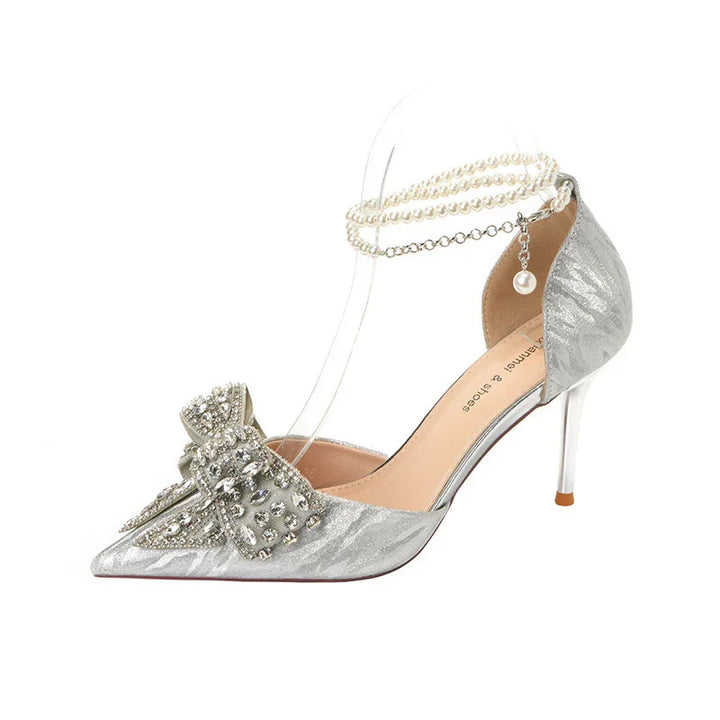 Silver high heel shoe with pointed toe, embellished bow, and pearl ankle strap